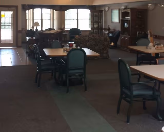 Interior view of a common area in a senior living facility with tables and chairs arranged for dining or socializing. There are large windows letting in natural light, a door with glass panels, and a seating area with a couch and armchairs in the background.