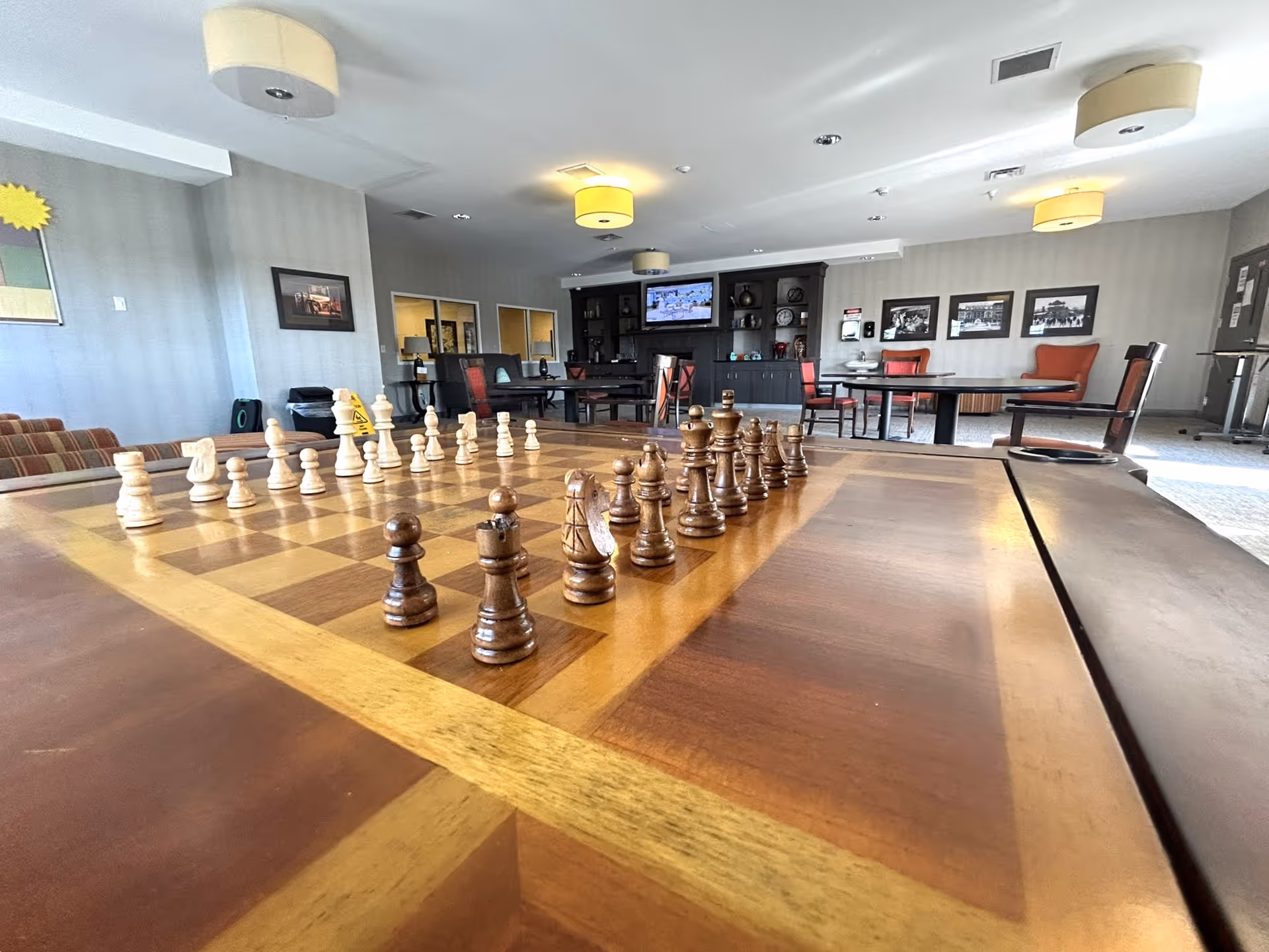 A spacious common room in a senior living facility with a large wooden chessboard in the foreground set up for a game. The room has several tables and chairs, framed pictures on the walls, a television mounted on a dark wooden cabinet, and soft ceiling lights. The atmosphere is calm and inviting.