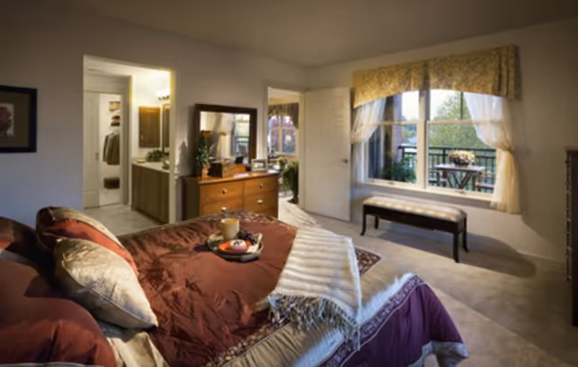 A cozy bedroom with a neatly made bed featuring burgundy and beige bedding. A tray with a cup and some food is placed on the bed. There is a wooden dresser with a mirror and decorative items, an open door leading to a bathroom, and a large window with sheer curtains and a valance, showing an outdoor patio with a table and chairs.