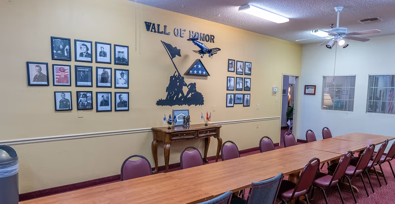A meeting or dining room with a long wooden table surrounded by red cushioned chairs. On the wall, there is a 'Wall of Honor' display featuring framed black and white photographs of military personnel, a silhouette of soldiers raising a flag, a folded American flag in a triangular case, and a model airplane hanging from the ceiling. The room has beige walls, a ceiling fan with lights, and windows with blinds.