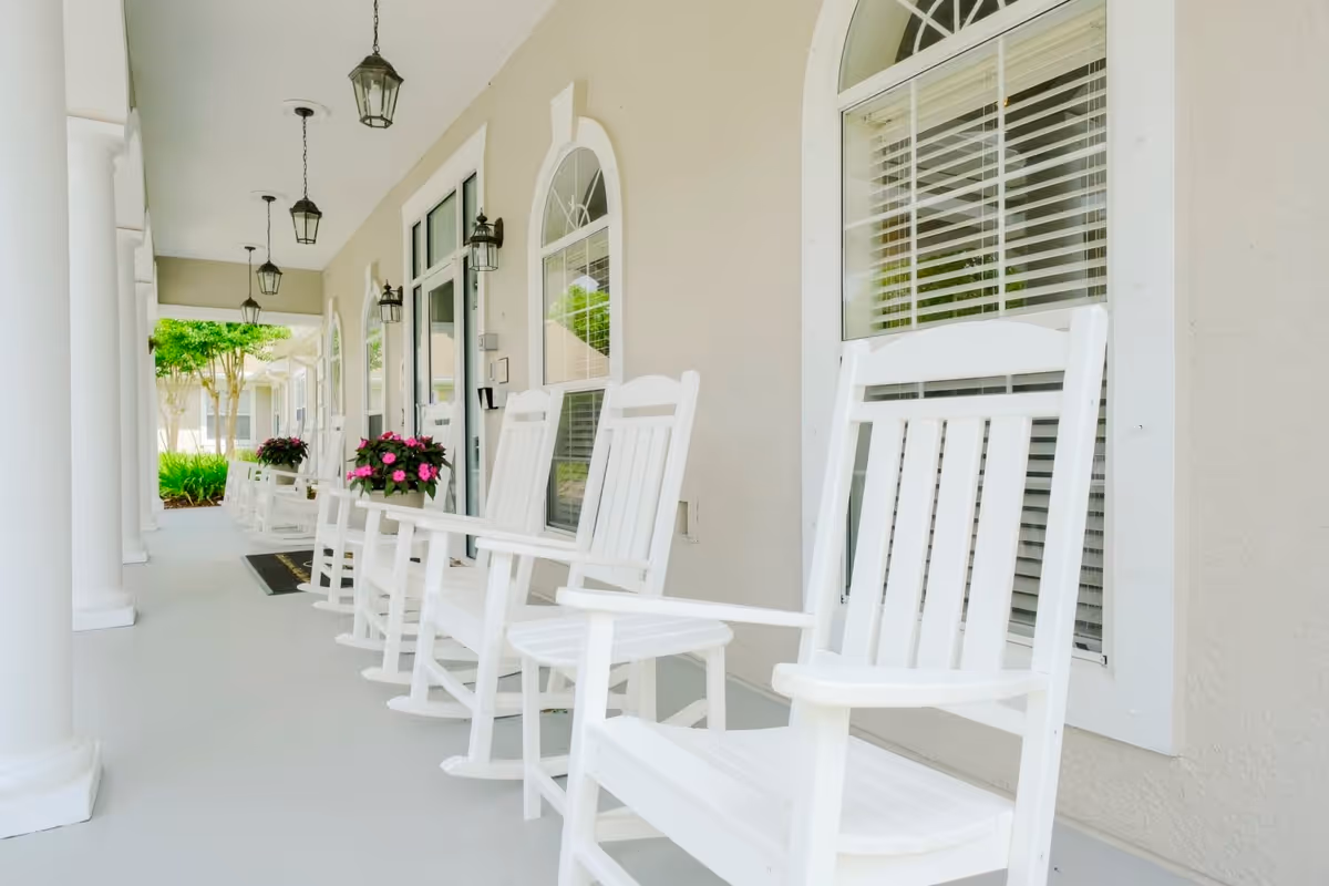 A covered porch area with white rocking chairs lined up along the wall of a beige building. There are hanging lantern-style lights on the ceiling and wall-mounted lanterns beside arched windows with white blinds. Potted pink flowers are placed on small tables between some of the chairs. White columns support the porch roof, and greenery is visible at the far end of the porch.