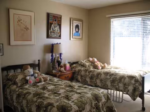 Two twin beds with palm-pattern spreads and stuffed animals in a shared senior living bedroom.