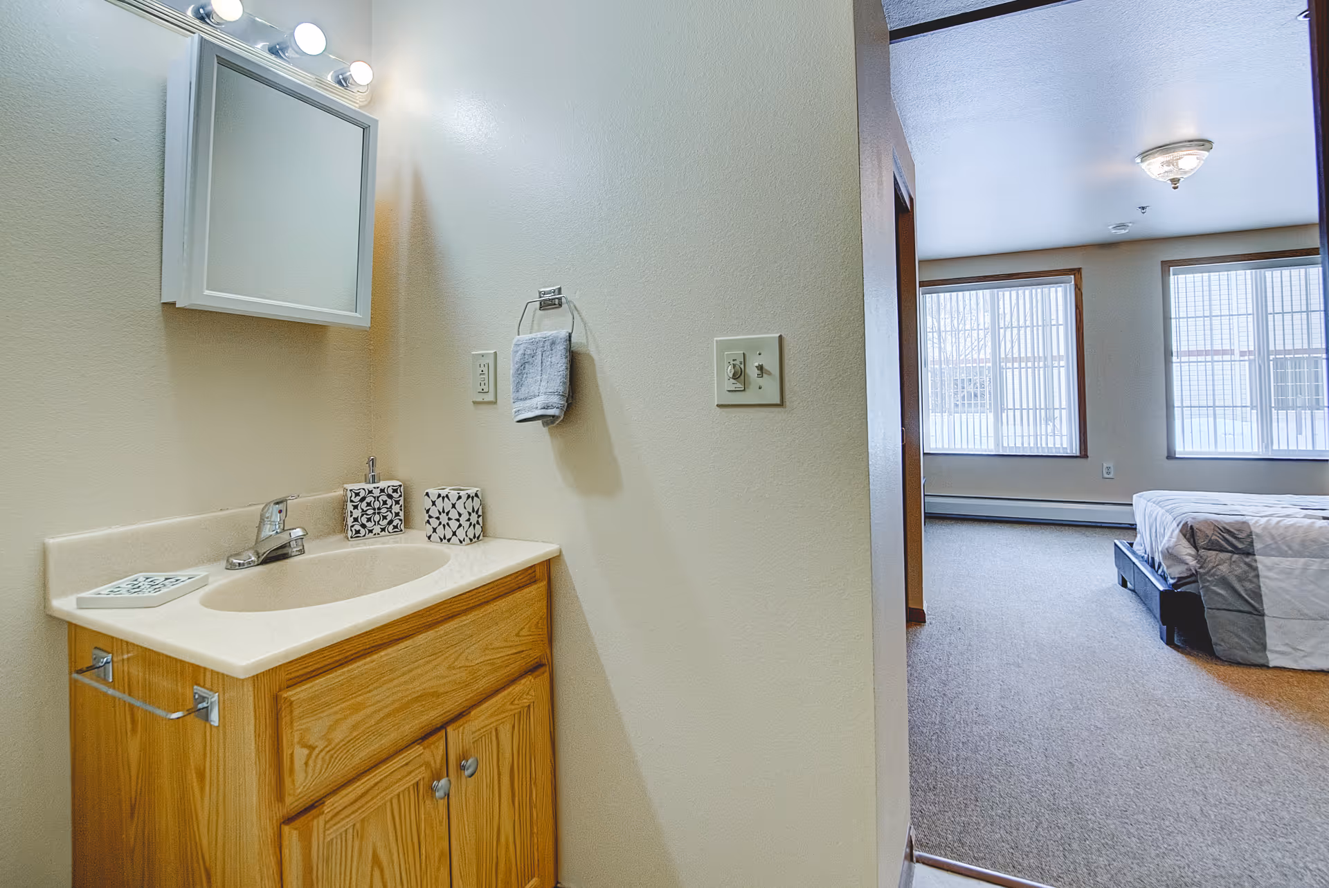View of a small bathroom sink area with a wooden cabinet, a mirror with lights above, a towel hanging on the wall, and decorative items on the countertop. The bathroom opens into a bedroom with a bed and large windows letting in natural light.