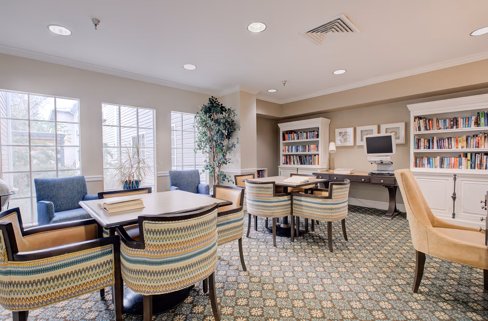 A bright and cozy reading room with large windows letting in natural light. The room features two tables surrounded by patterned and leather chairs, two blue armchairs near the windows, and two white bookshelves filled with books. A computer monitor is placed on a dark wooden desk against the wall, and there are framed pictures hanging above it. The floor has a patterned carpet, and there is a tall potted plant near the corner.