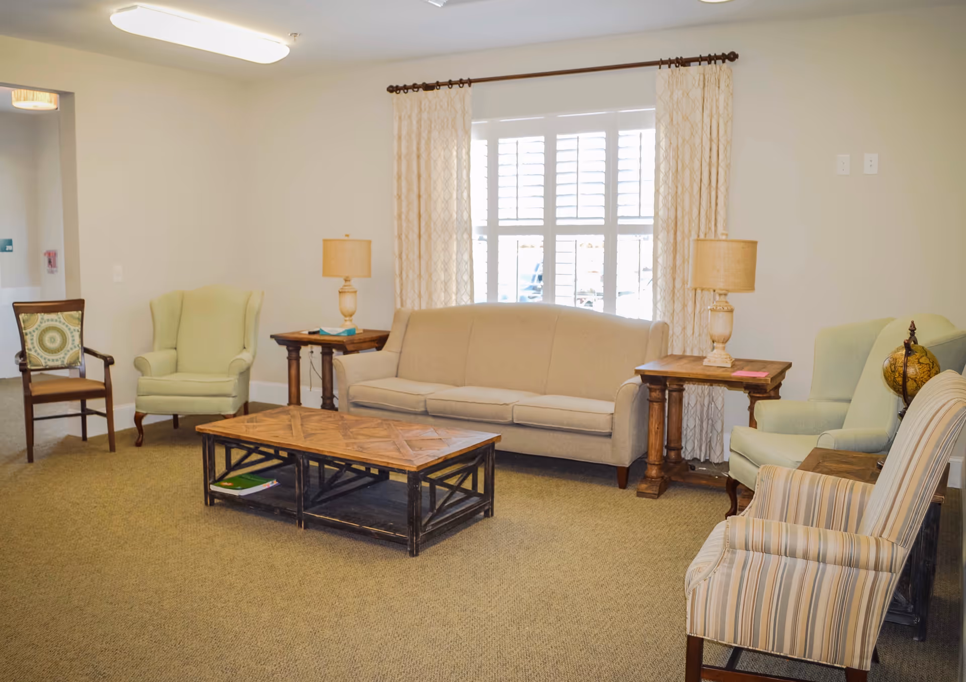Well-lit communal living room with a sofa, several armchairs, a wooden coffee table and side tables with lamps in front of a window.