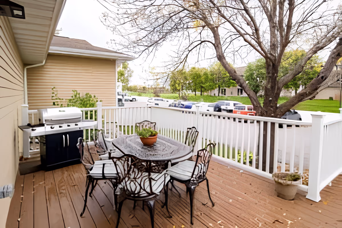 Outdoor patio area with a wooden deck, a metal table with six matching chairs, a potted plant on the table, a barbecue grill against the house wall, and a large tree nearby. In the background, there are several parked cars and green grassy areas.