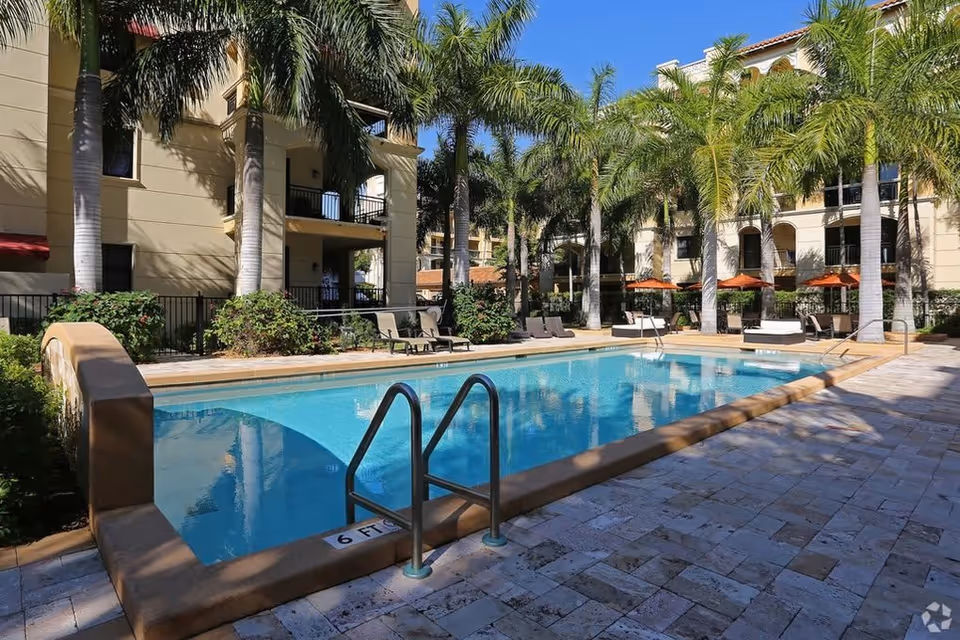 Outdoor swimming pool surrounded by palm trees and lounge chairs with umbrellas, adjacent to a multi-story residential building under a clear blue sky.
