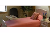 A single bed with a pink bedspread and decorative pillow in a small furnished room with a chair, side table, and potted plant.