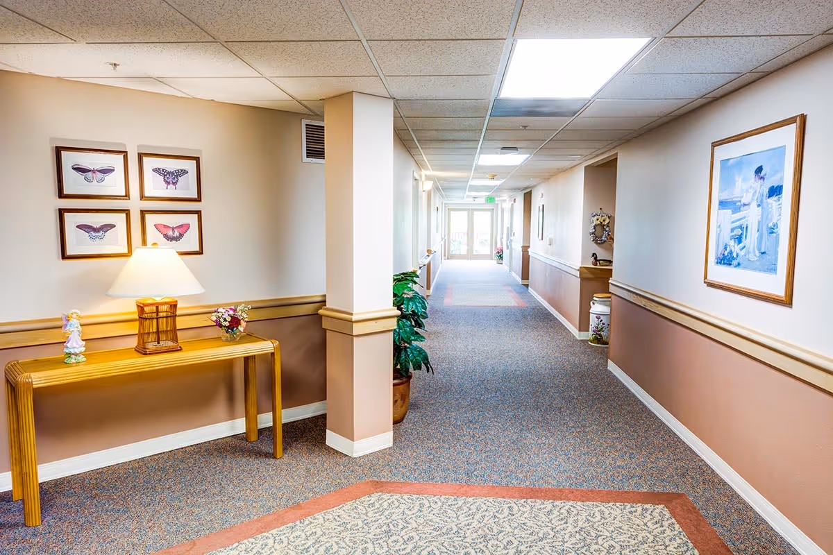 Carpeted interior hallway of a senior living facility lined with artwork, a console table with a lamp, and potted plants leading to glass doors.