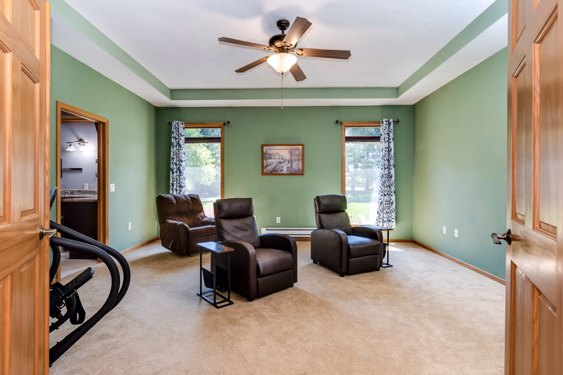 Sunlit living room with green walls featuring three recliners, side tables, a ceiling fan, and two windows with patterned curtains.