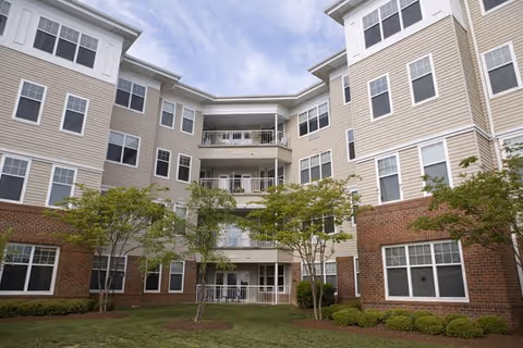 Exterior courtyard view of a multi-story senior living building with balconies, many windows, and landscaped grass and small trees.