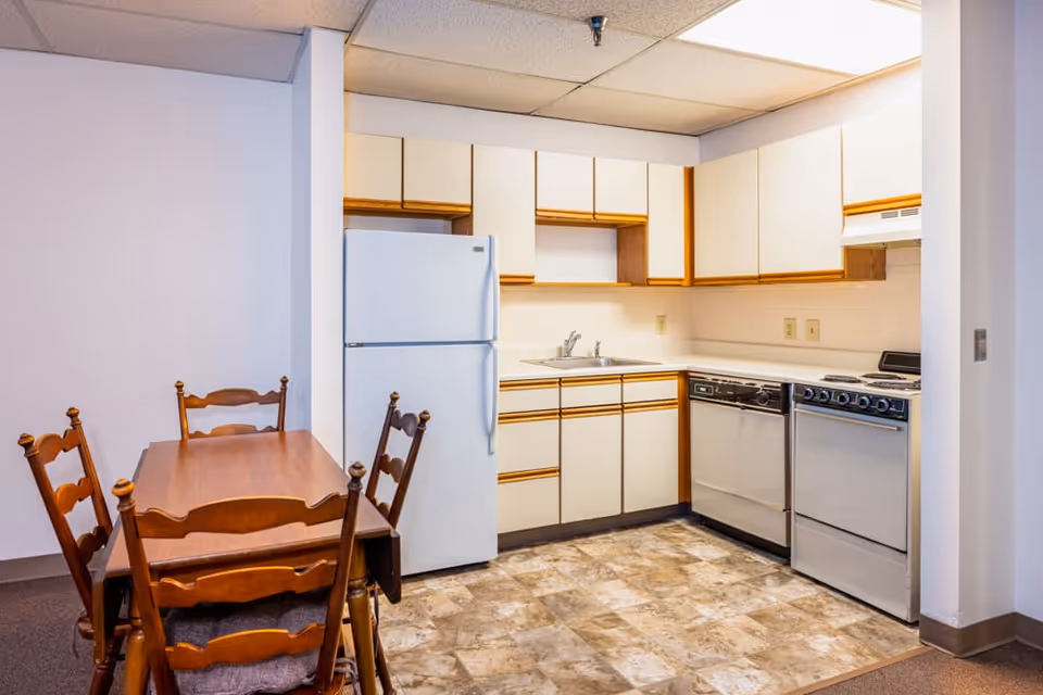 A small kitchen area with white cabinets and wooden trim, a white refrigerator, a dishwasher, a stove, and a double sink. Adjacent to the kitchen is a wooden dining table with four matching chairs. The floor has a patterned linoleum surface, and the walls are painted white.