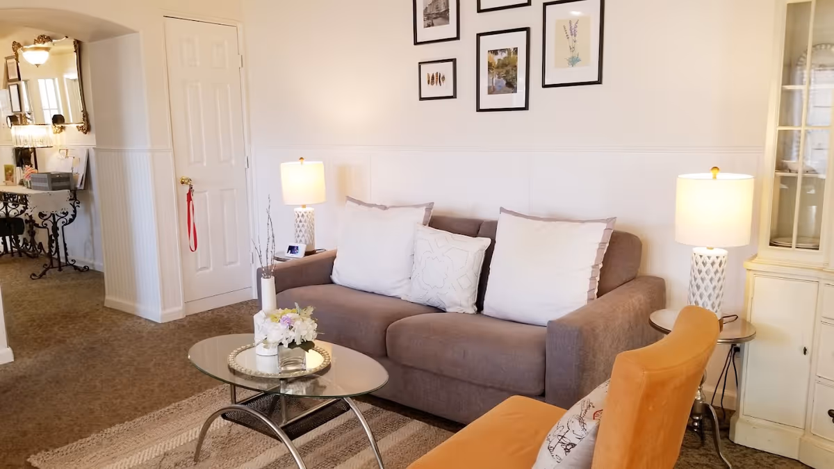 A cozy living room with a gray sofa adorned with three white pillows, a glass coffee table with a floral arrangement, an orange armchair with a decorative pillow, two table lamps on either side of the sofa, framed artwork on the wall, and a carpeted floor.
