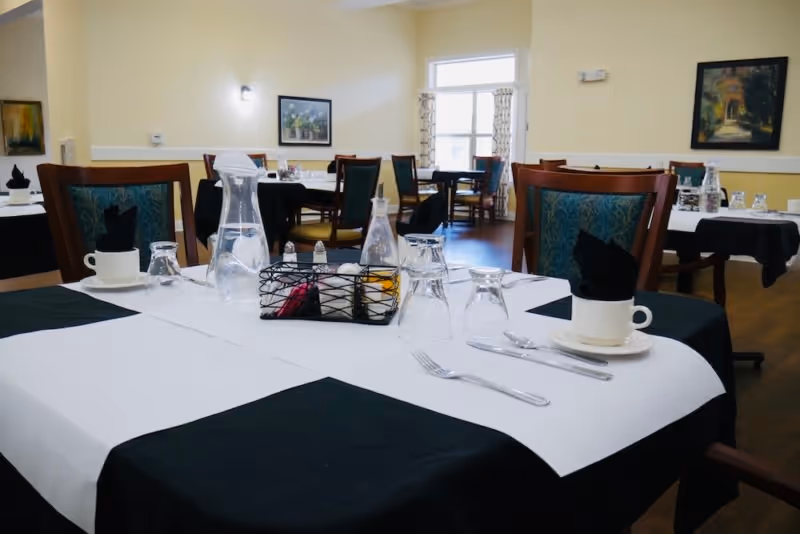 A dining room in a senior living facility with tables set for a meal. Each table is covered with a white tablecloth and black placemats, with glassware, silverware, cups, and napkins neatly arranged. The room has wooden chairs with patterned upholstery, light yellow walls, framed artwork, and windows letting in natural light.