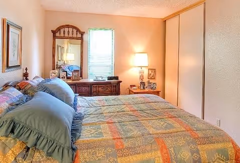 Sunlit bedroom with a patterned bedspread, dresser with mirror, nightstand lamp, and sliding closet doors.