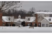 Snow-covered front exterior of a single-story brick and siding building with a central cupola, chimneys, and leafless trees in the background under a cloudy sky.