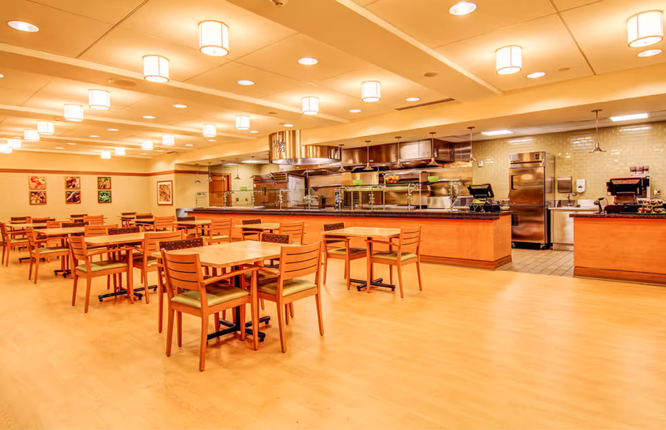 A spacious dining area with multiple wooden tables and chairs arranged neatly on a light wood floor. The room is well-lit with ceiling lights and features a serving counter with stainless steel kitchen equipment and appliances in the background. The walls have framed pictures of fruits and vegetables.