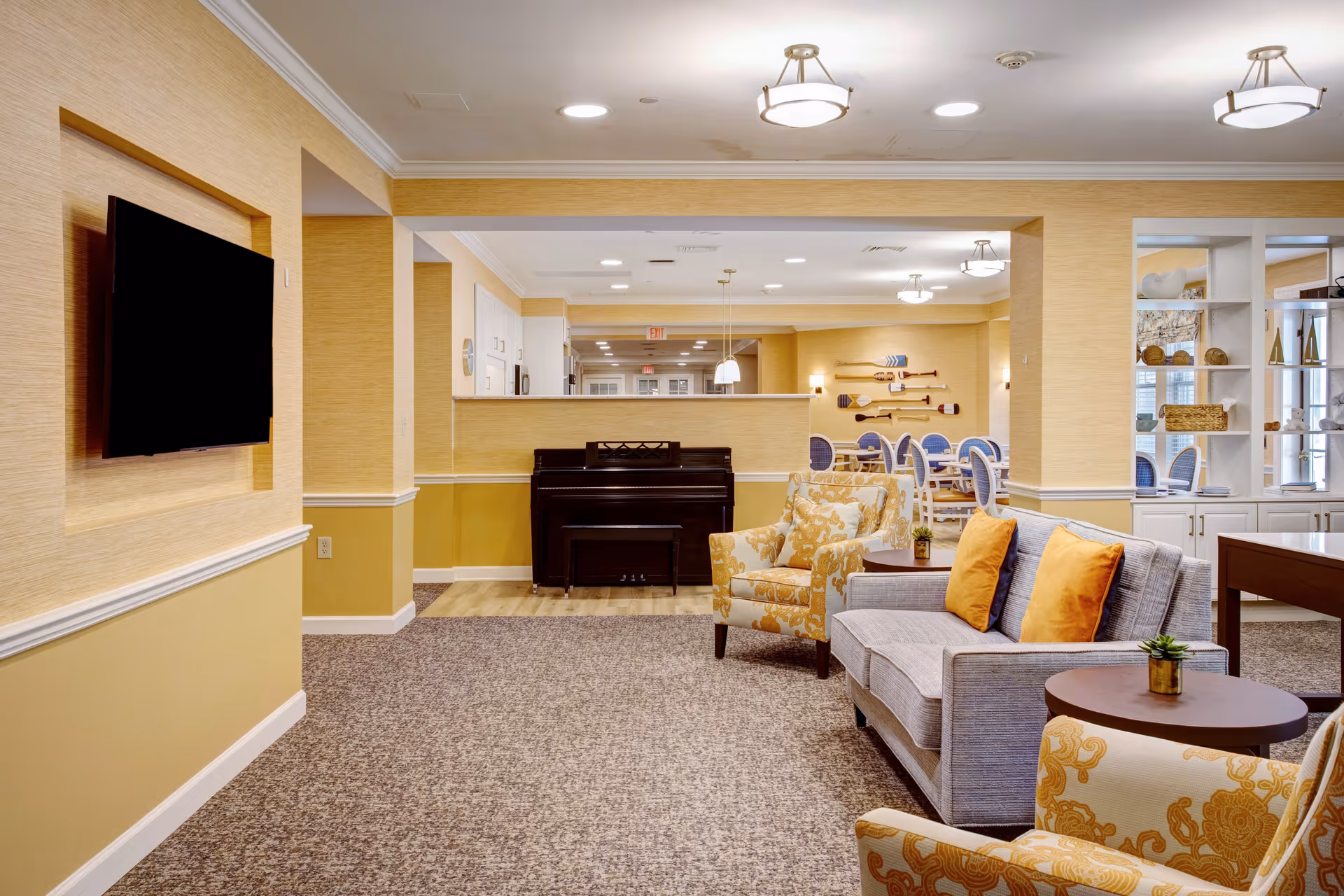 A cozy senior living facility common area with a gray sofa adorned with orange pillows, two yellow patterned armchairs, a round wooden side table with a small plant, a black piano against a half wall, and a wall-mounted flat-screen TV. In the background, there is a dining area with blue cushioned chairs and decorative paddles mounted on the wall. The room features warm yellow walls, carpeted flooring, and ceiling lights.