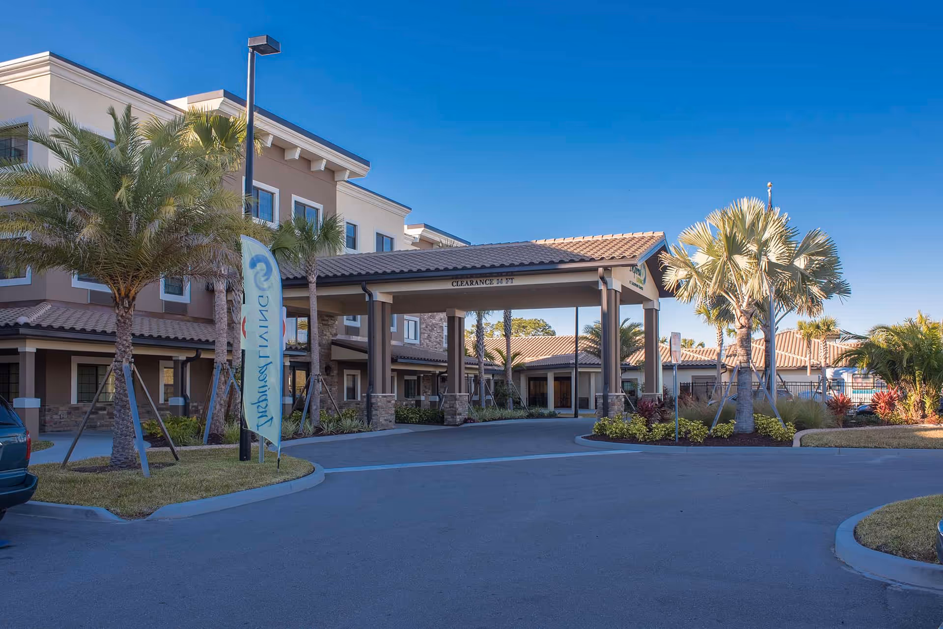 Entrance and porte-cochère of a two-story senior living building with palm trees and a circular driveway.