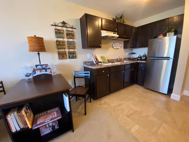 Small kitchenette and dining area with dark wood cabinets, a stainless-steel refrigerator, countertop appliances, a table with a lamp, and a bookshelf.