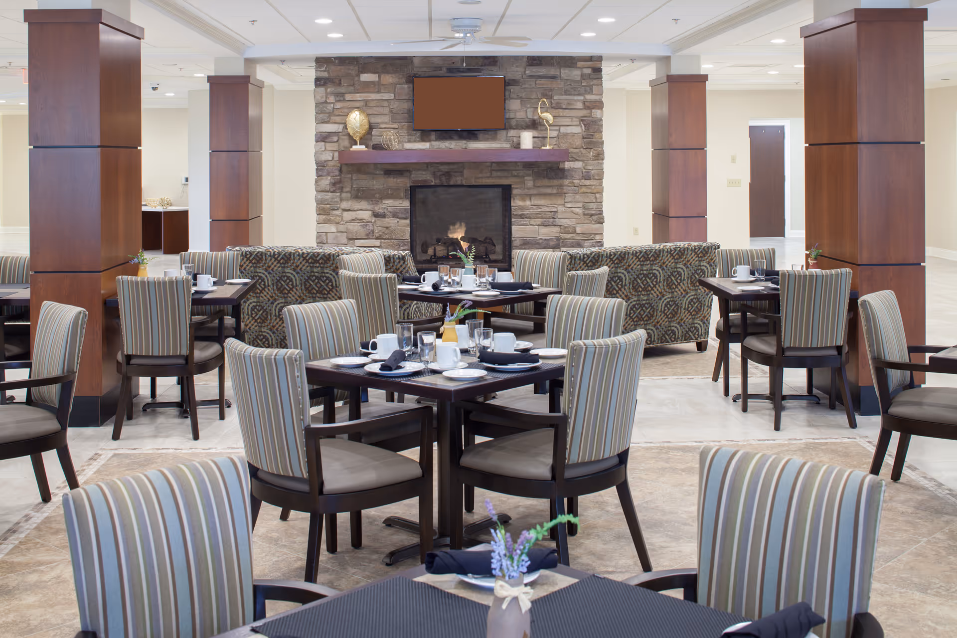 A dining area in a senior living facility with multiple tables set for meals, featuring striped upholstered chairs and a stone fireplace with a mounted TV above it. The room has wooden columns and patterned sofas in the background.