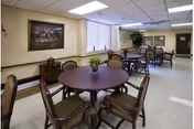 Bright communal dining room with round wooden tables, upholstered chairs, plants, and framed artwork on the walls.