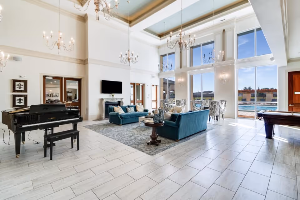 Spacious and elegant living room area with high ceilings and large windows letting in natural light. The room features two teal sofas with cushions arranged around a coffee table on a patterned rug. A black grand piano with a matching bench is on the left side, and a pool table is visible on the right. Chandeliers hang from the ceiling, and a flat-screen TV is mounted above a fireplace. Outside the windows, a swimming pool and outdoor seating area can be seen.
