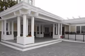 Exterior view of a single-story building with a covered entrance supported by white columns and a paved driveway area in front. The building has large windows and a modern design with white walls and a gray roof.