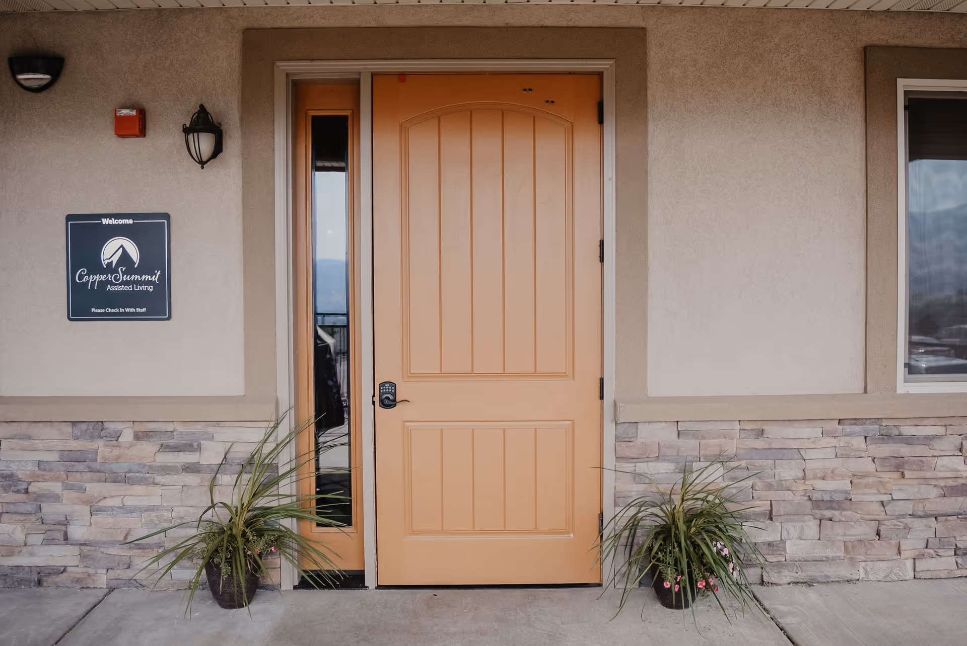 Front entrance of Copper Summit Assisted Living with a tan door, narrow sidelight window, potted plants, and a wall sign.