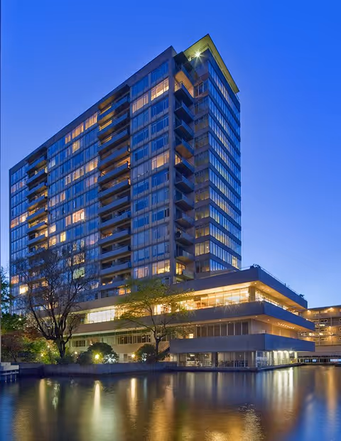 A tall modern senior living community building at dusk with many windows illuminated, reflected in a calm body of water in the foreground, surrounded by trees and landscaping.
