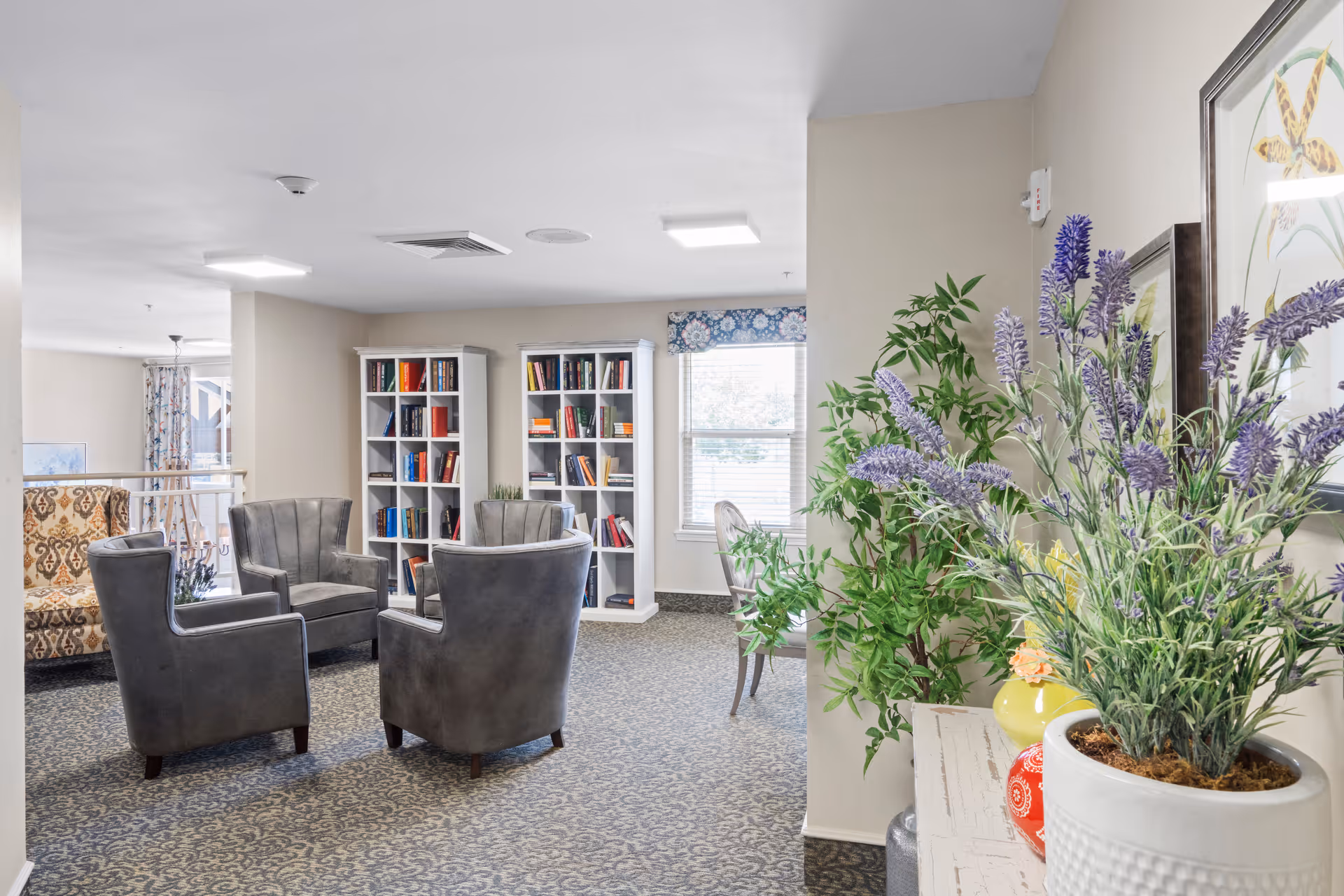 A cozy sitting area in a senior living facility with four gray armchairs arranged around a small table, two white bookshelves filled with books against the wall, a window with a floral valance, and decorative plants including a large pot of purple flowers in the foreground.