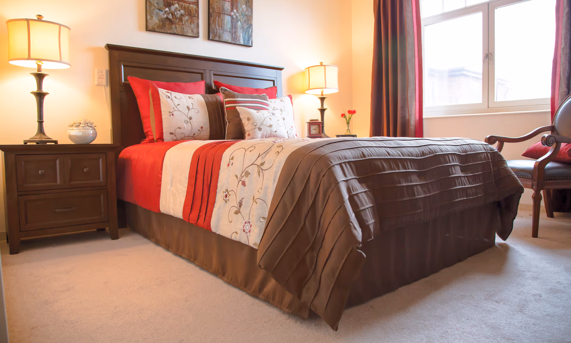 Well-lit bedroom with a made bed dressed in red and brown bedding, nightstands with lamps, and a window.
