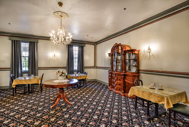 Elegantly furnished dining room with small set tables, a central round table, chandelier, and a wooden china cabinet.