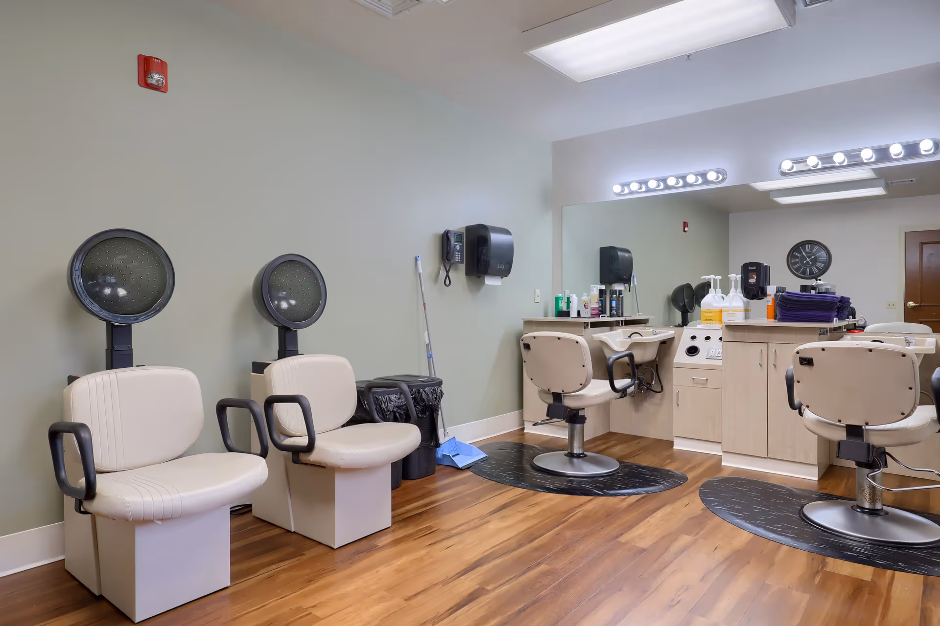 Interior of a senior living facility hair salon with two beige hair drying chairs on the left and three beige salon chairs in front of mirrors on the right. The room has wooden flooring, light green walls, and various hair care products on the counters.
