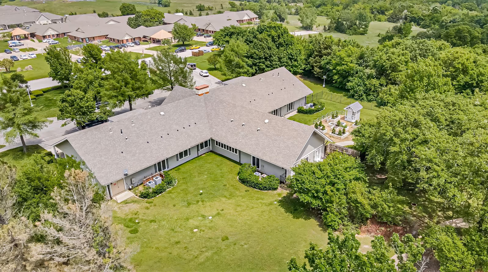 Aerial view of a senior living facility named The Homestead of Denison, showing a large building with a gray roof surrounded by green lawns, trees, and a garden area. Several cars are parked near other buildings in the background.