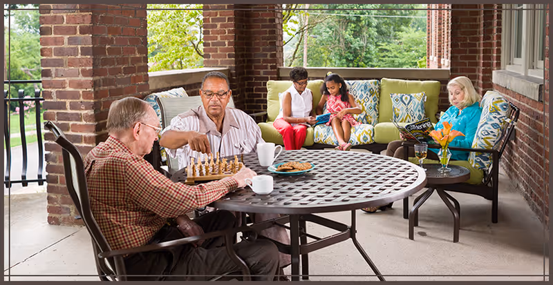 Two elderly men playing chess at a round metal table on a covered patio with brick pillars. In the background, a woman and a young girl sit on a cushioned outdoor sofa looking at a tablet, while another elderly woman reads a magazine on a chair. The setting is bright and surrounded by greenery.