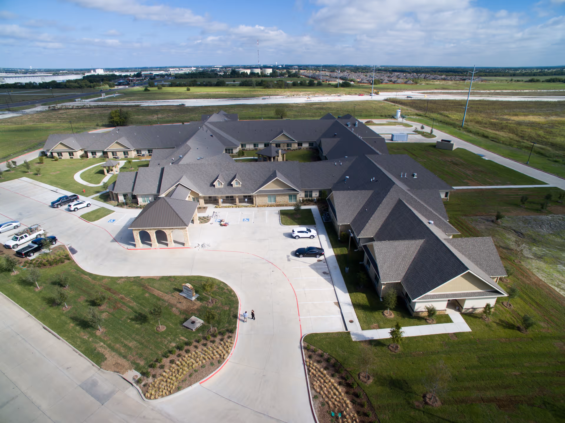 Aerial view of a large single-story healthcare and rehabilitation facility with multiple wings, surrounded by green lawns and parking areas with several cars. The building has a gray roof and beige exterior walls. The surrounding area includes open fields and a distant view of other buildings and infrastructure.