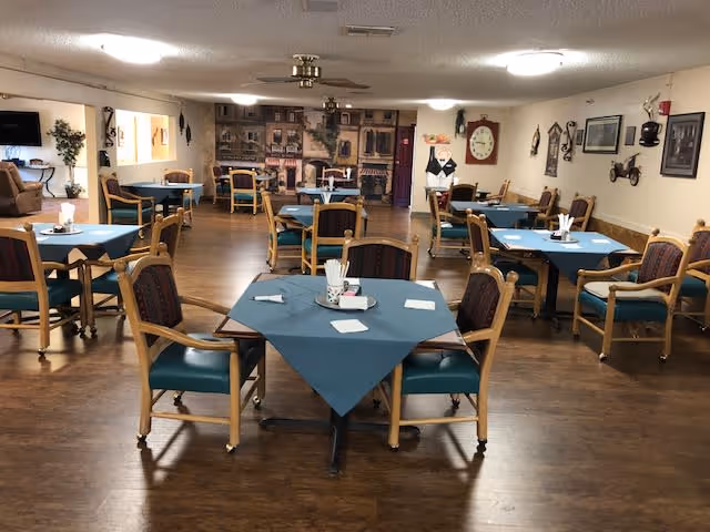 A dining room with multiple tables covered with blue tablecloths, each surrounded by wooden chairs with green cushions. The room has wooden flooring, ceiling lights, and wall decorations including framed pictures and a large clock. There is a mural on the back wall depicting a street scene with buildings.