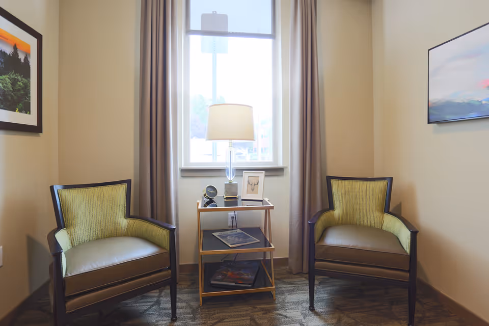 A cozy seating area with two armchairs upholstered in green and brown fabric, positioned on either side of a small gold and glass side table. The table holds a lamp, a small clock, and a framed picture. Behind the table is a window with beige curtains, and the walls are decorated with framed artwork.