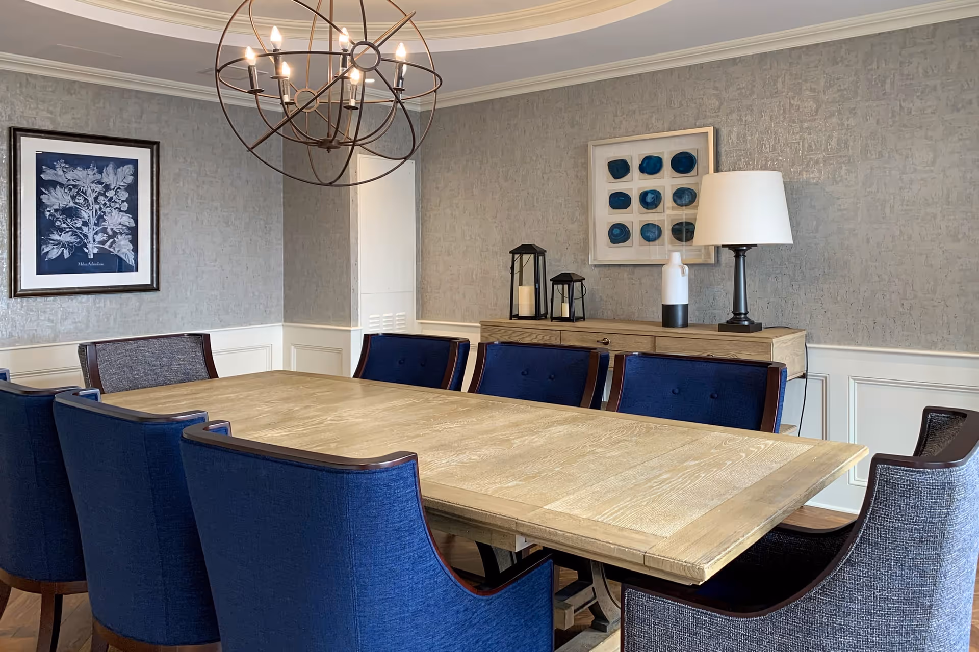 A dining room with a large wooden table surrounded by eight upholstered chairs, six in blue fabric and two in gray fabric. The room features a modern spherical chandelier with exposed bulbs hanging above the table. On the wall behind the table is a wooden sideboard with decorative items including two black lanterns, a white and black vase, and a table lamp with a white shade. The walls are decorated with two framed artworks, one depicting botanical prints and the other abstract blue shapes.