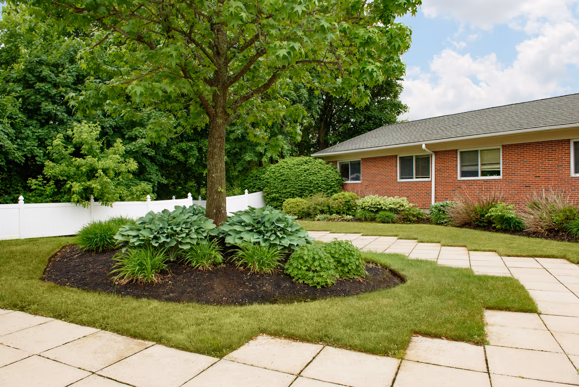 A landscaped outdoor area at Life Care Center of Leominster featuring a tree surrounded by various green plants and shrubs, a white fence in the background, a brick building with windows, and a paved walkway under a partly cloudy sky.