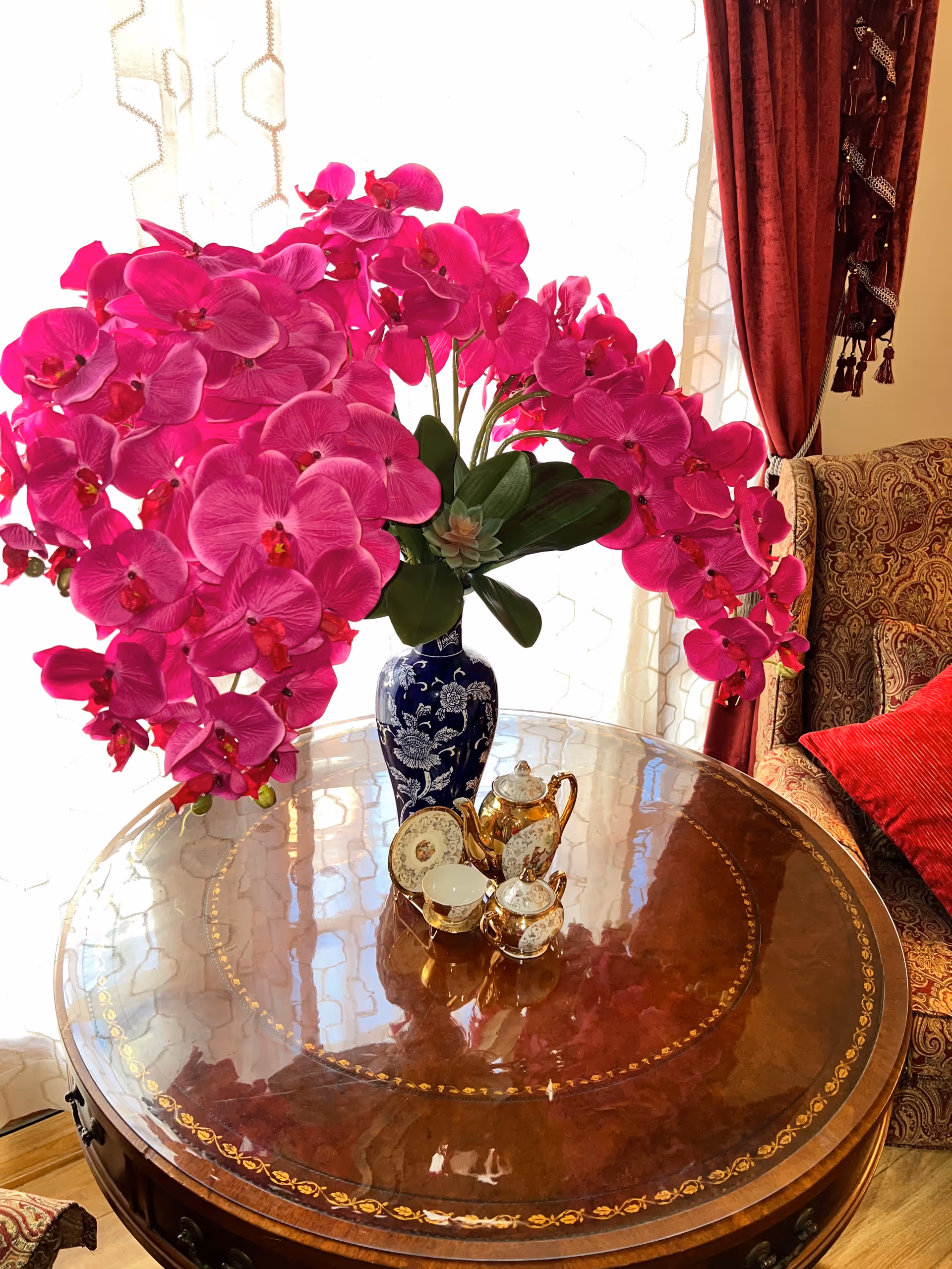 A round wooden table with intricate gold detailing on the surface, holding a blue and white vase filled with vibrant pink orchids. Next to the vase is a small ornate tea set with a teapot, sugar bowl, creamer, and a cup and saucer. In the background, there is a patterned armchair with a red cushion and red curtains with tassels, and sheer white curtains letting in natural light.