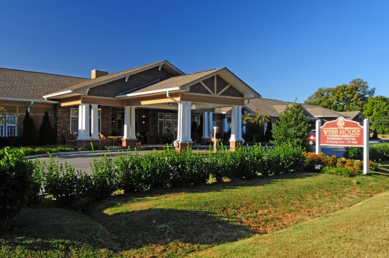 Exterior view of Webb House Retirement Center building with a covered entrance supported by white columns, surrounded by green bushes and a well-maintained lawn under a clear blue sky.