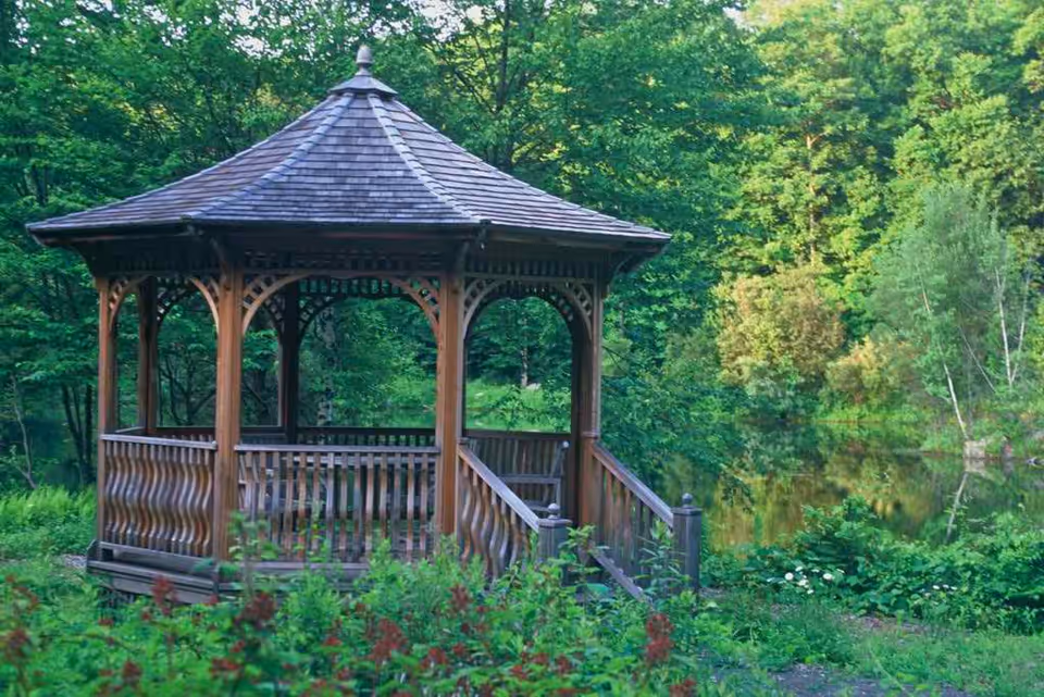 A wooden gazebo with a shingled roof situated in a lush green garden area near a small pond surrounded by trees and various plants.