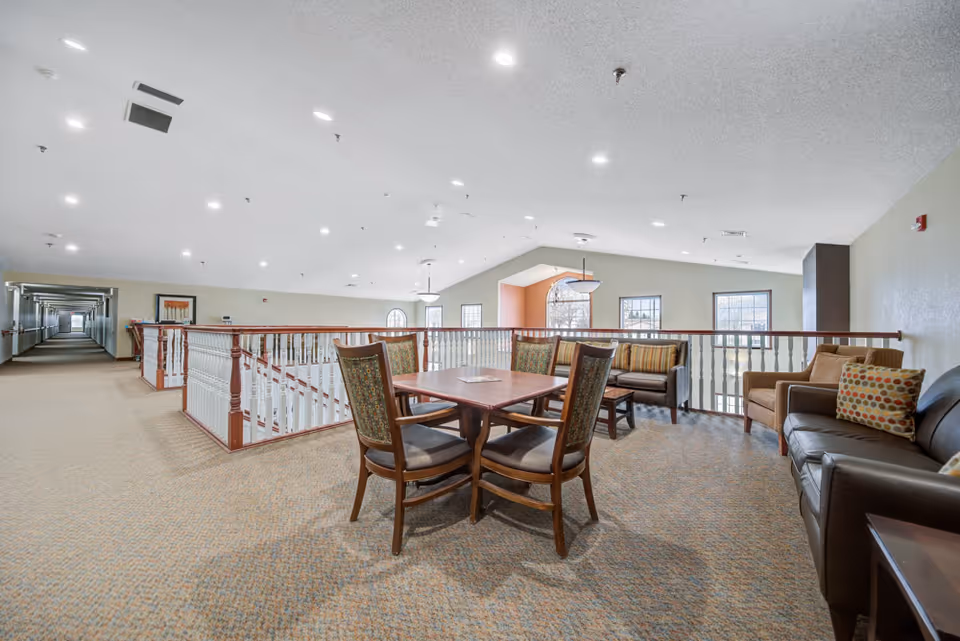 A spacious and well-lit common area in a senior living facility featuring a wooden table with four chairs in the center, surrounded by several cushioned sofas with patterned pillows. The area overlooks a stairwell with white railings and has large windows allowing natural light to fill the space. The ceiling is high with recessed lighting, and a long hallway extends from the left side of the room.