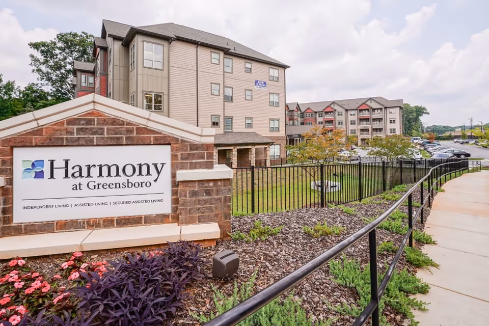 Exterior view of Harmony at Greensboro senior living facility showing a brick sign with the facility name and description, a landscaped area with flowers and shrubs, a black metal fence, a sidewalk, and multi-story residential buildings in the background under a partly cloudy sky.