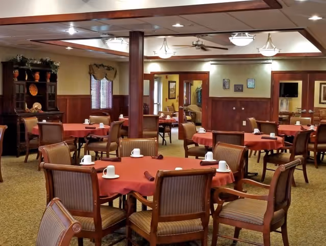 A dining room in a senior living facility with multiple round tables covered with red tablecloths. Each table is surrounded by wooden chairs with cushioned seats and backs. White coffee cups and rolled napkins are placed on the tables. The room has warm lighting, wood paneling on the walls, and decorative elements including a cabinet with plates and plants, as well as framed pictures on the walls.