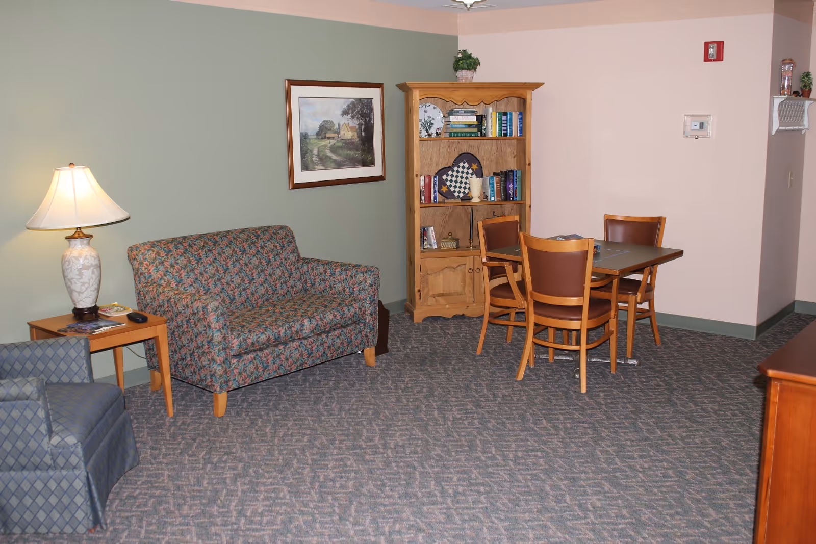 A cozy living area with a floral patterned loveseat, a blue armchair, a wooden side table with a lamp, a wooden bookshelf filled with books and decorative items, and a small dining table with four chairs. The walls are painted in soft green and beige tones, and there is a framed landscape painting on the wall.