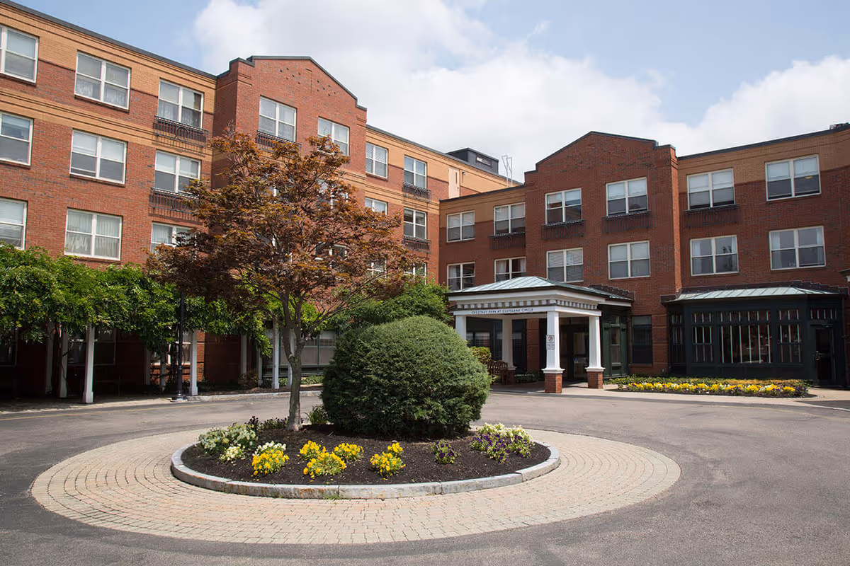 Exterior view of Chestnut Park at Cleveland Circle, a multi-story brick building with multiple windows. In front of the building is a circular driveway with a landscaped roundabout featuring a tree, a large bush, and colorful flowers. The entrance has a covered portico with white columns and a sign above it.