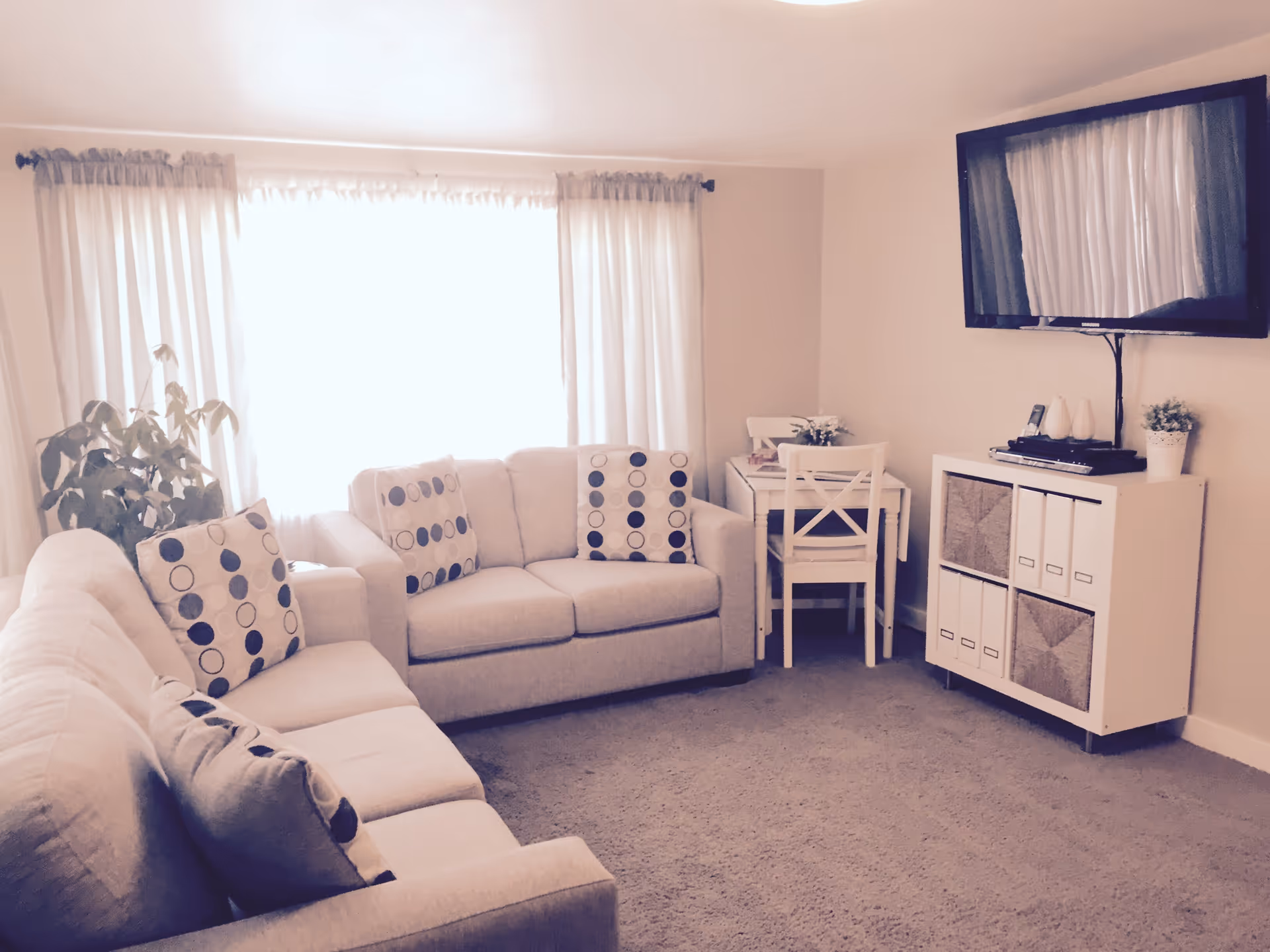 A cozy living room with two beige sofas adorned with patterned cushions, a large window with sheer curtains letting in bright light, a small white table with two chairs in the corner, a white cabinet with storage boxes and decorative items, and a flat-screen TV mounted on the wall.